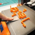 Dłoń z łopatką zdejmuje pomarańczowy wydruk 3D z platformy drukarki