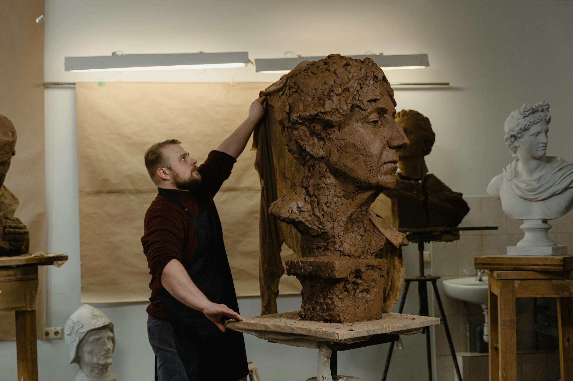 Rzeźbiarz w pracowni modeluje dużą glinianą głowę otoczony rzeźbami